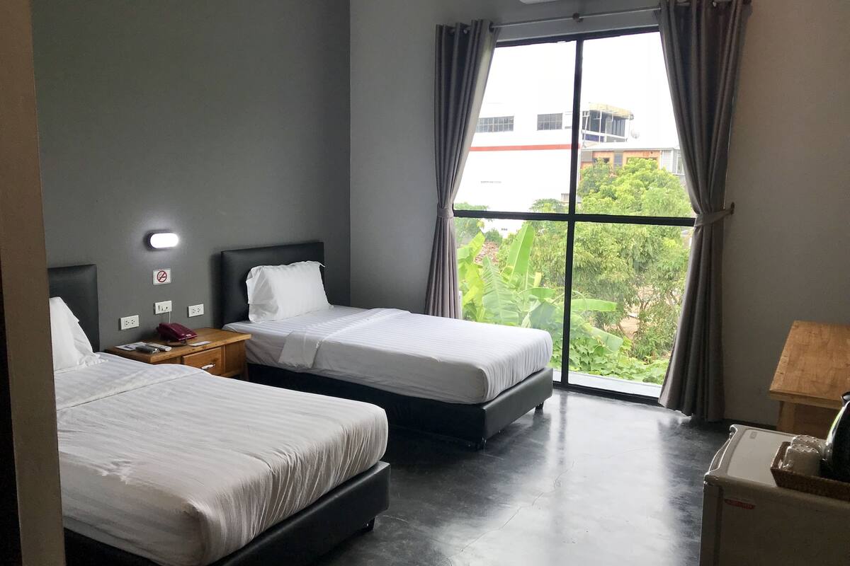 A hotel room with two beds, a nightstand, a lamp, and a view of greenery and buildings outside.