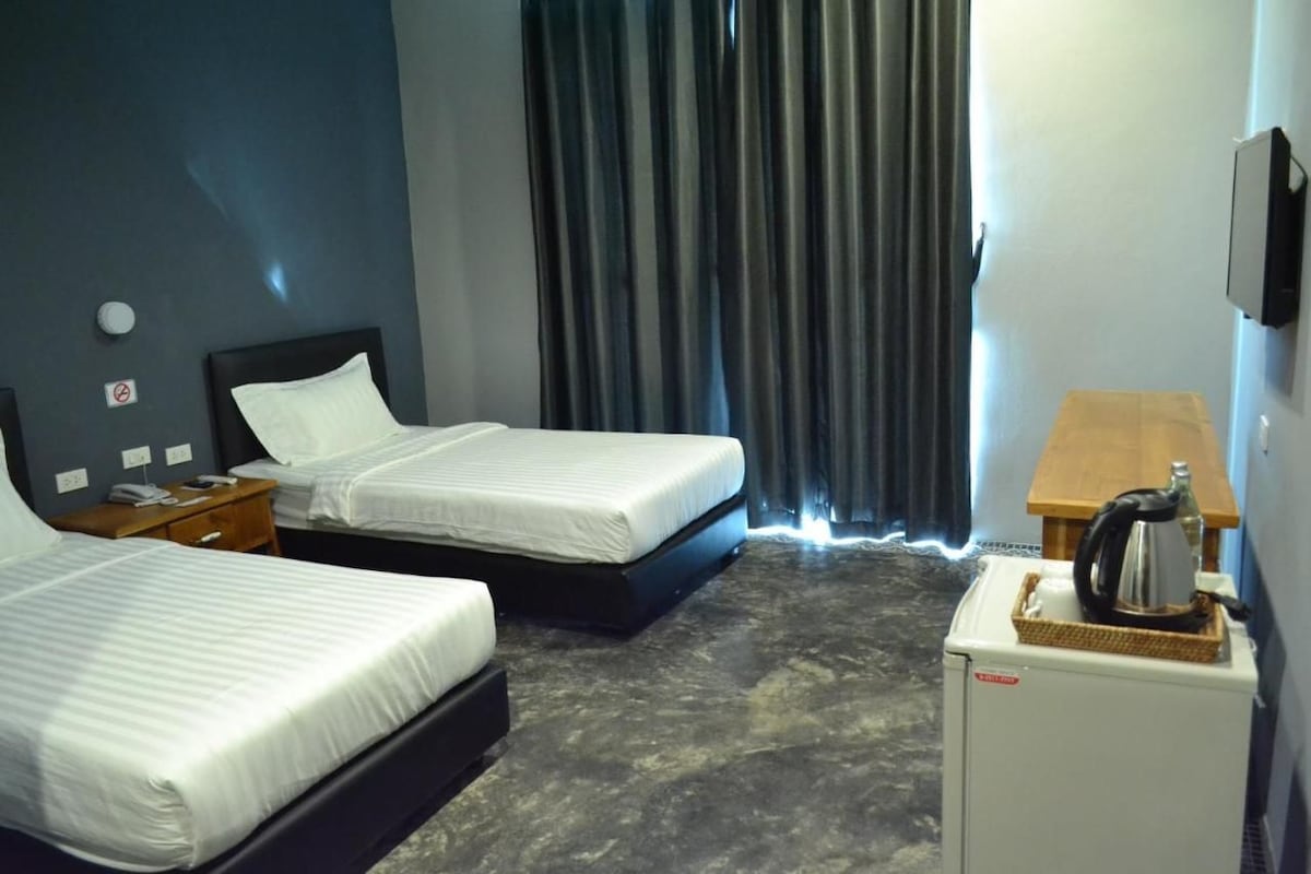 A hotel room with two beds, a small table with a kettle, and a refrigerator.