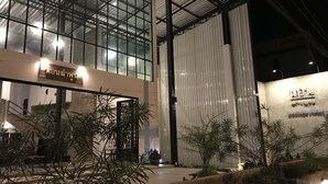 Un edificio moderno con fachada de vidrio, un letrero con escritura tailandesa y un jardín cuidado con plantas en macetas.
