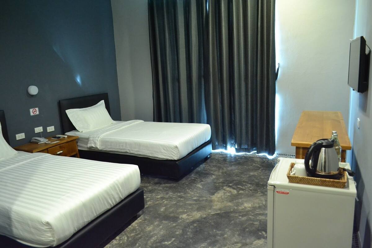 A hotel room with two beds, a small table with a kettle, a refrigerator, and a window with curtains.