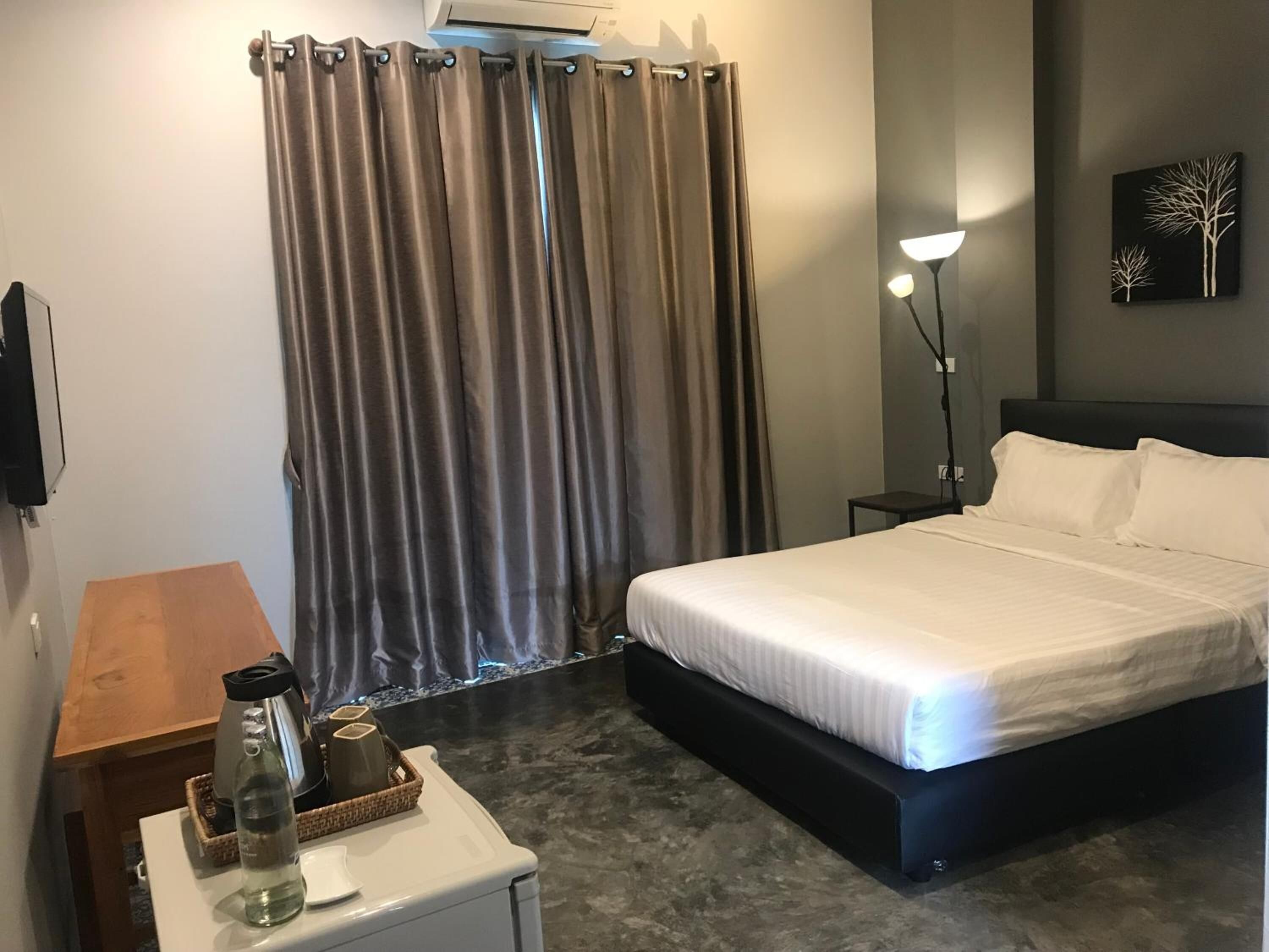 A hotel room with a bed, a bedside table with a lamp, a small table with a water bottle and a basket, and a wall-mounted air conditioner.