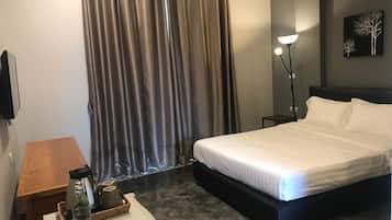A hotel room with a bed, a bedside table with a lamp, a small table with a water bottle and a basket, and a wall-mounted air conditioner.