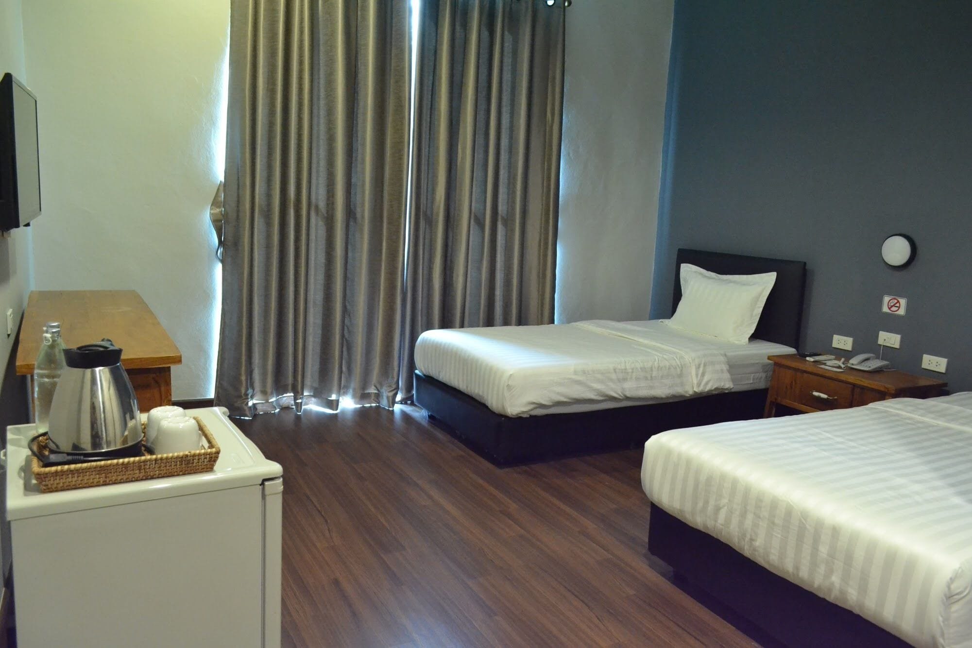 Quarto de hotel com duas camas, uma mesa pequena com chaleira, mesa de cabeceira com telefone e relógio na parede.