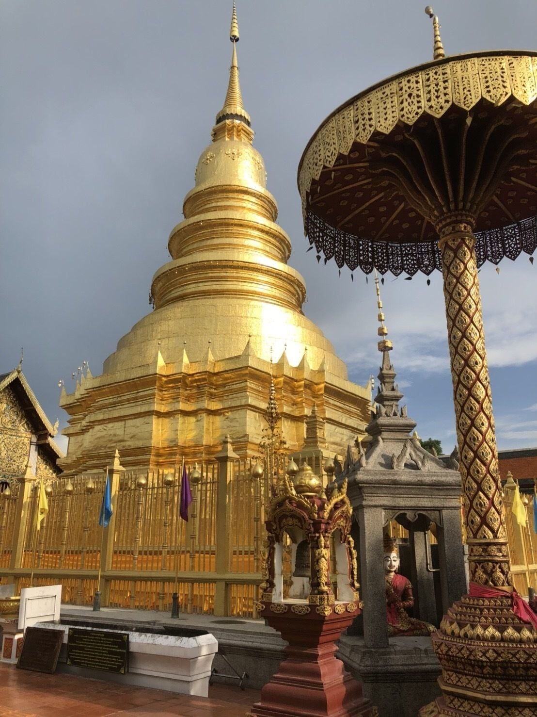 A large golden stupa with intricate carvings and a tiered structure, surrounded by traditional architecture and a decorative umbrella.