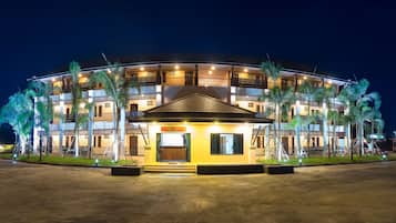 Un hotel de varios pisos con un patio central, que cuenta con una entrada bien iluminada y palmeras alrededor.