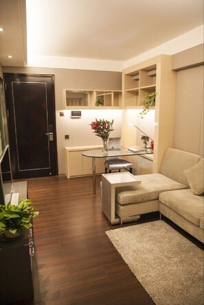 Un espacio habitable pequeño con un sofá, una mesita y una mesa de centro de vidrio. Se observa una puerta de madera oscura y una planta a la izquierda.