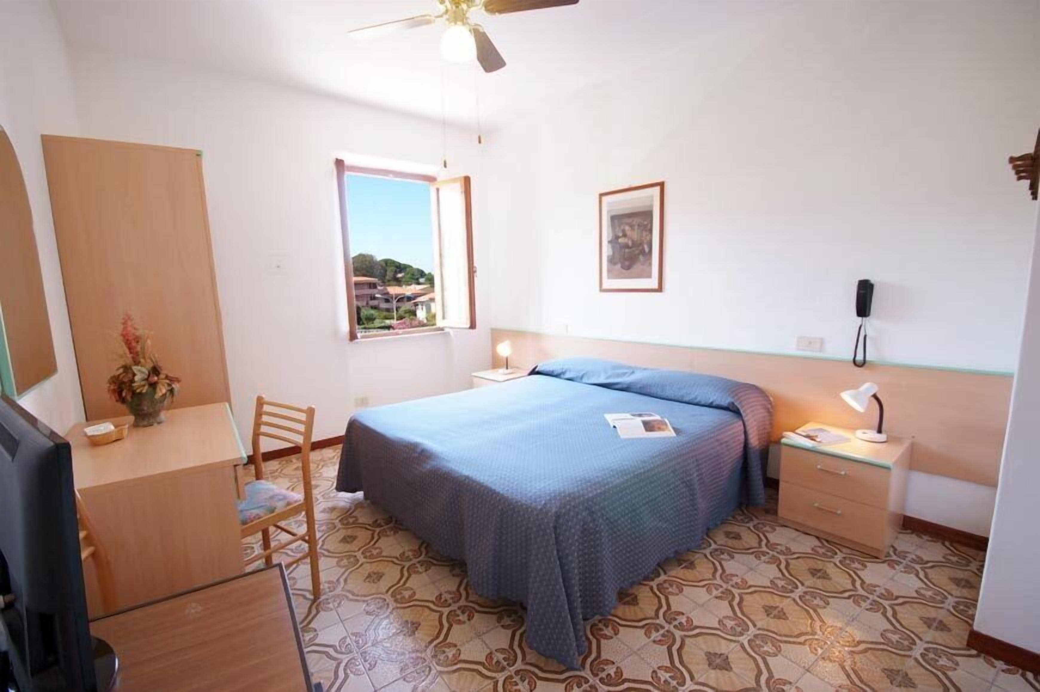 A bedroom with a bed, a desk, a chair, a nightstand, a lamp, and a window with a view.