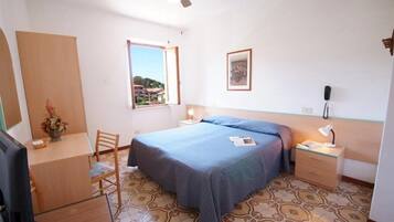 A bedroom with a bed, a desk, a chair, a nightstand, a lamp, and a window with a view.