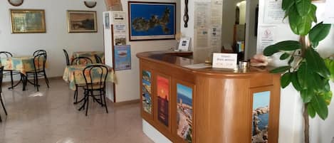A reception area with a wooden counter, framed pictures, and a map on the wall. Tables and chairs are arranged for dining.