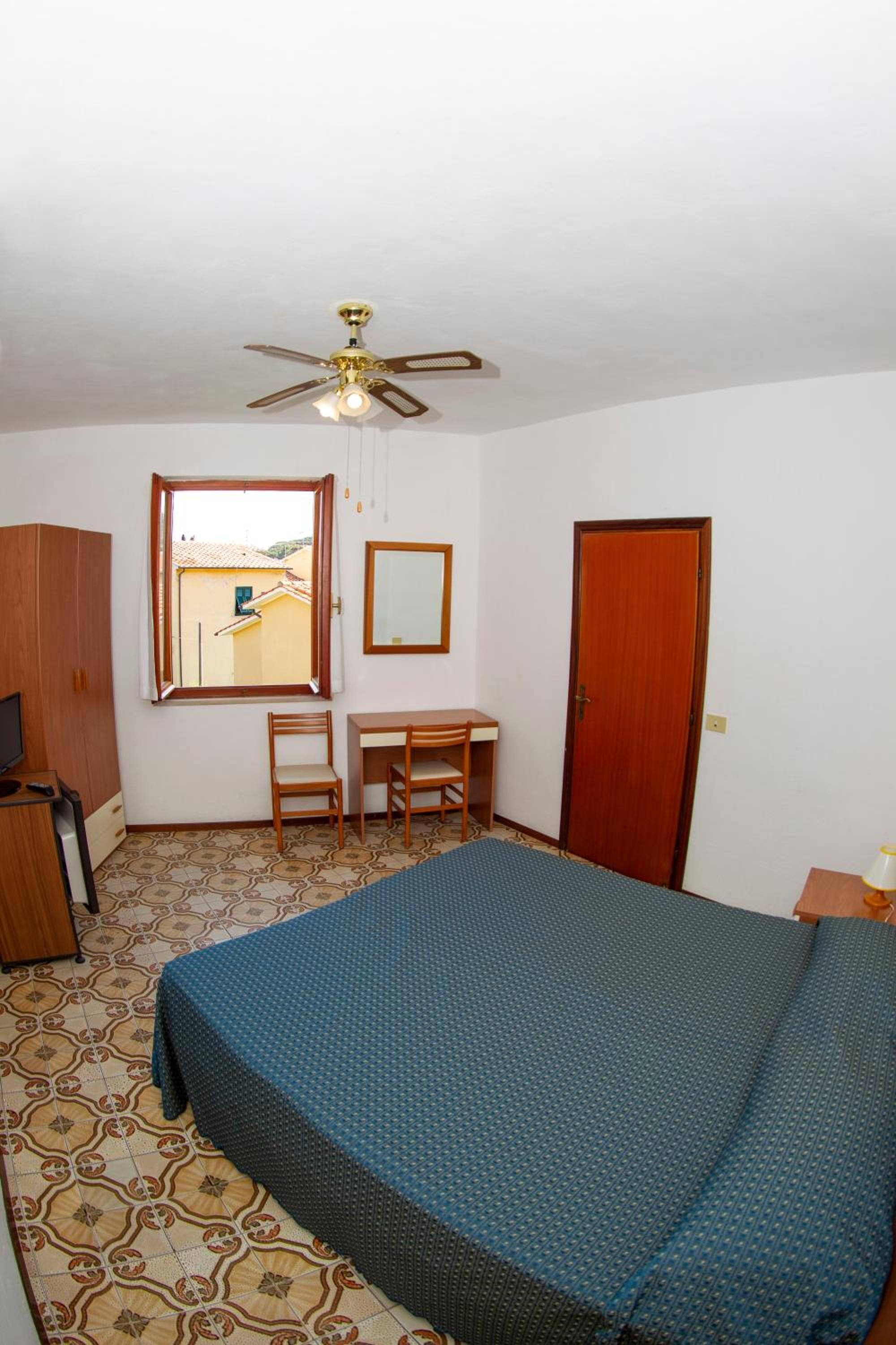 A bedroom with a bed, a desk, a chair, a wardrobe, a ceiling fan, a window, and a door.
