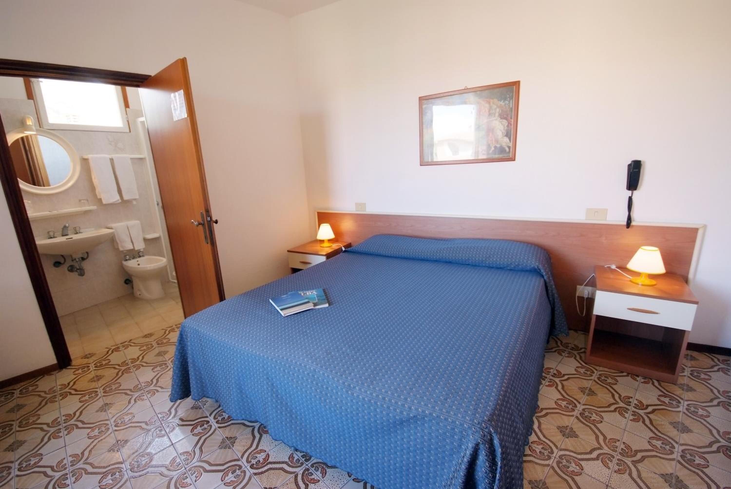 A hotel room with a bed, two bedside tables with lamps, a bedside phone, a painting on the wall, and a bathroom visible through an open door.
