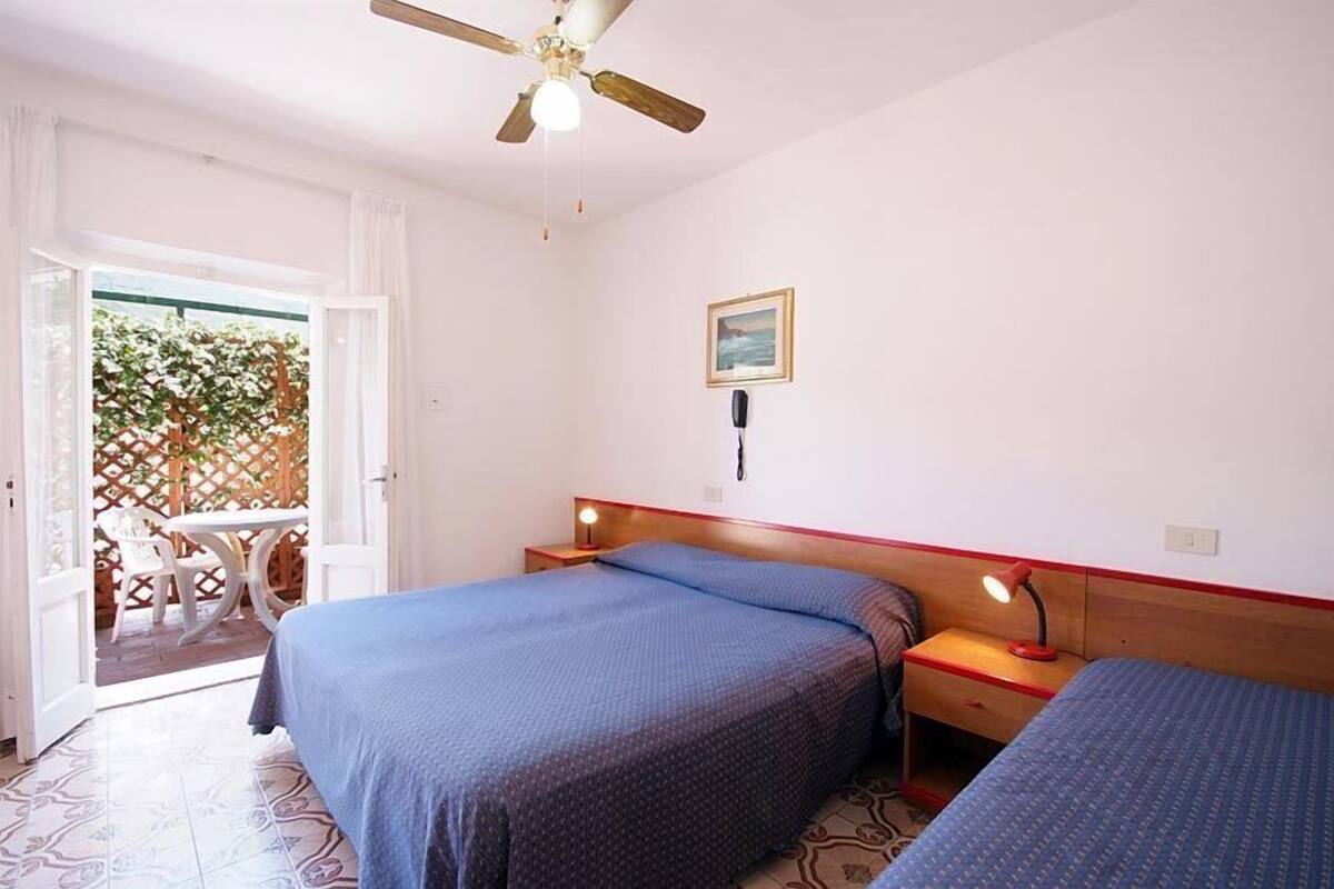 Une chambre avec deux lits, un ventilateur de plafond, une table de chevet avec une lampe et une fenêtre donnant sur un paysage verdoyant.