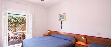 A bedroom with two beds, a ceiling fan, a bedside table with a lamp, and a window with a view of greenery outside.