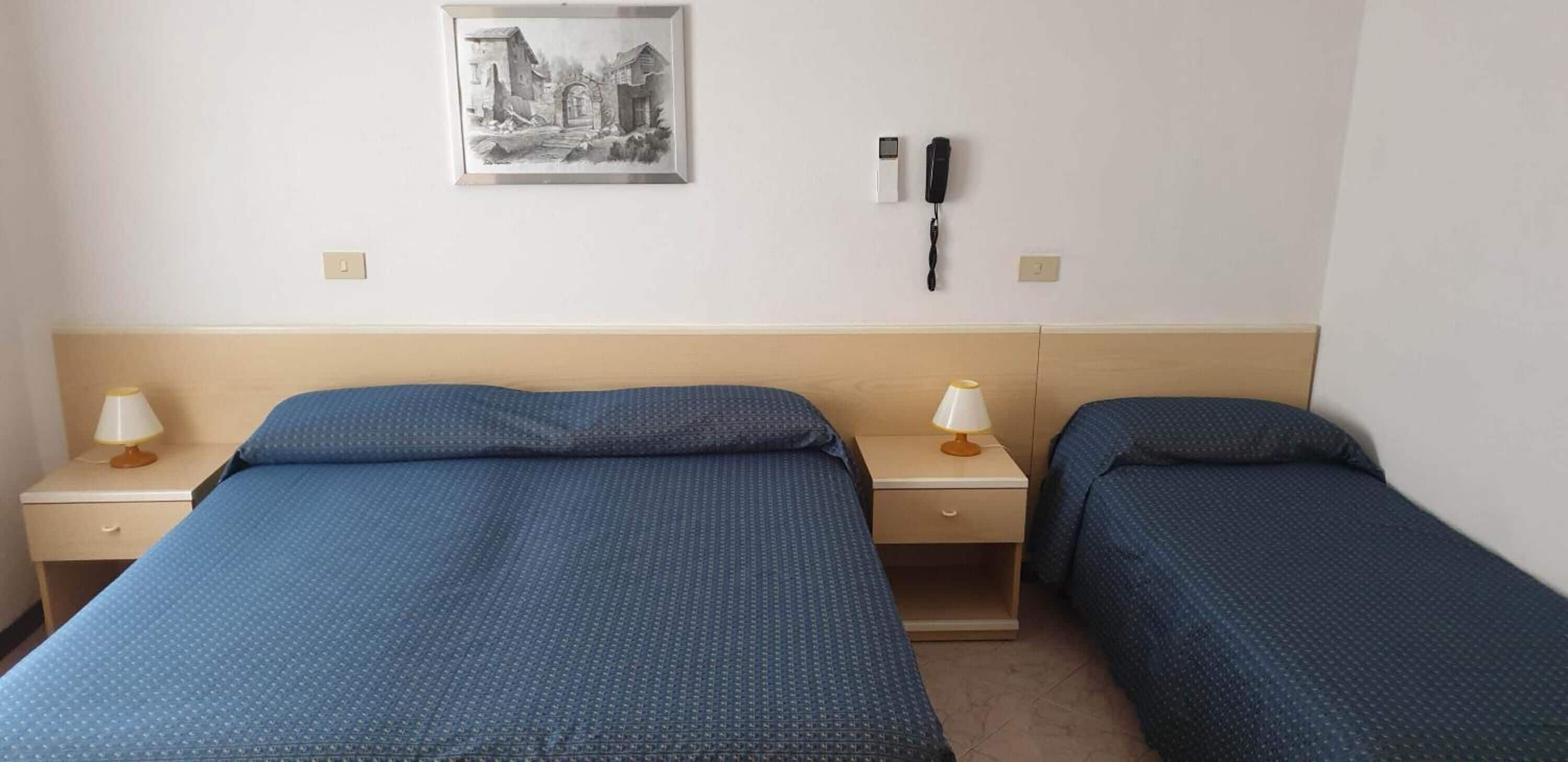 Camera d'albergo con due letti, un comodino con lampada, un telefono e un quadro appeso al muro.
