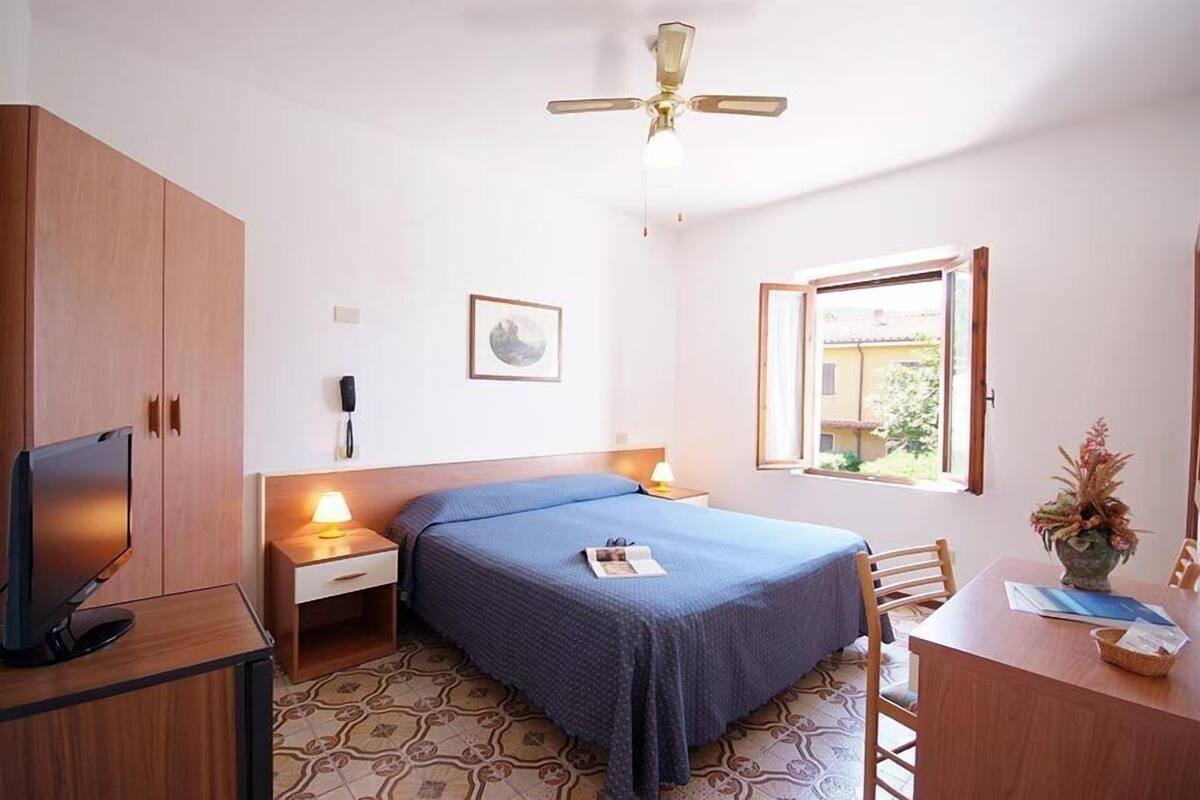 Une chambre à coucher comprenant un lit, un bureau, une télévision, un ventilateur de plafond, une fenêtre et un téléphone fixé au mur.