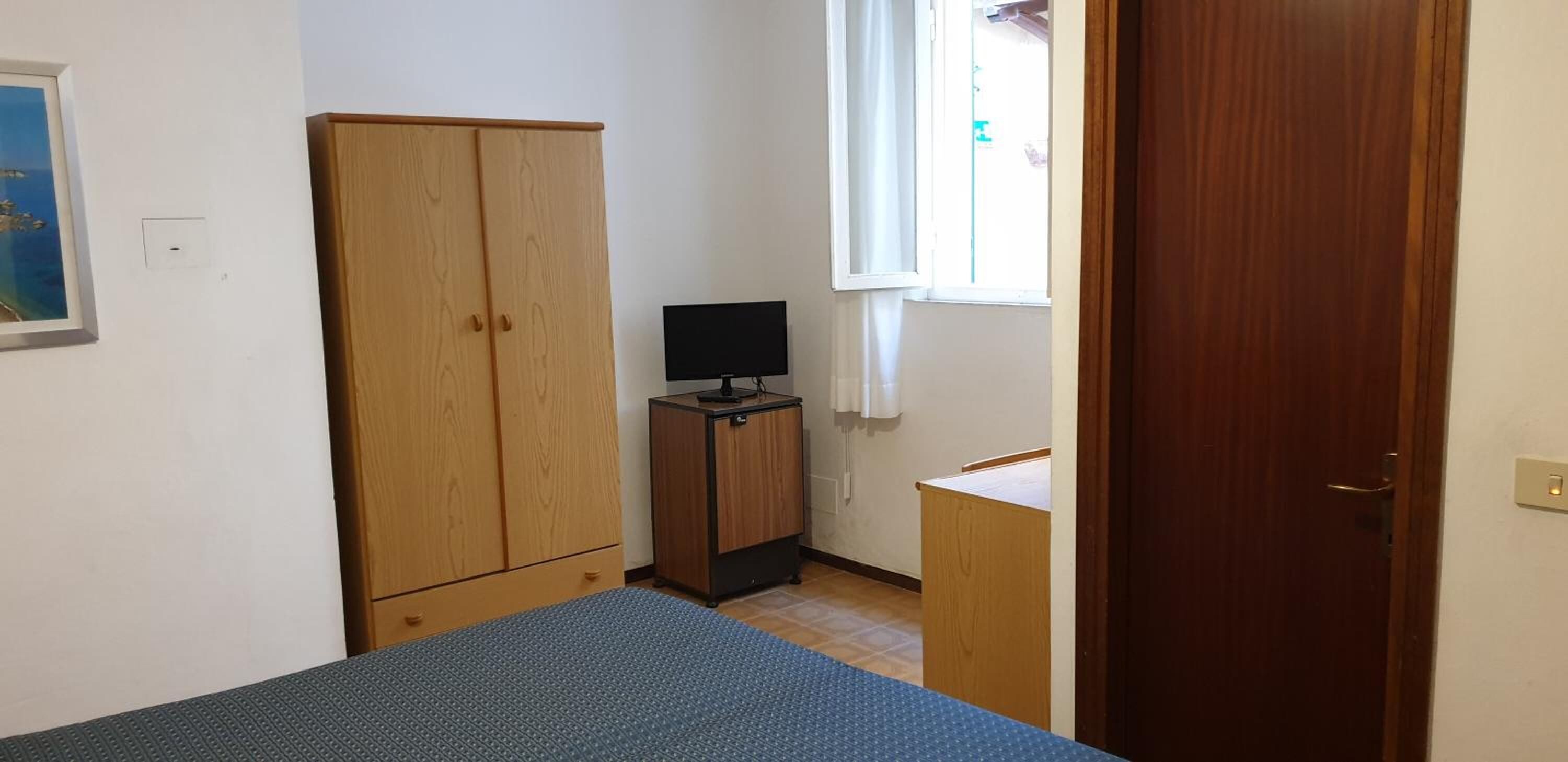 A small room with a bed, a wooden wardrobe, a small cabinet with a TV, and a window with curtains.