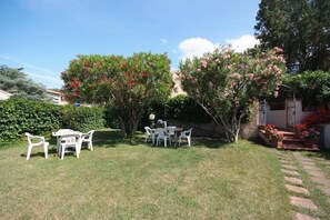 A backyard with a small patio area, a table, and chairs. There are flowering trees and a hedge in the background. - Hotel Anselmi (Marciana Marina)