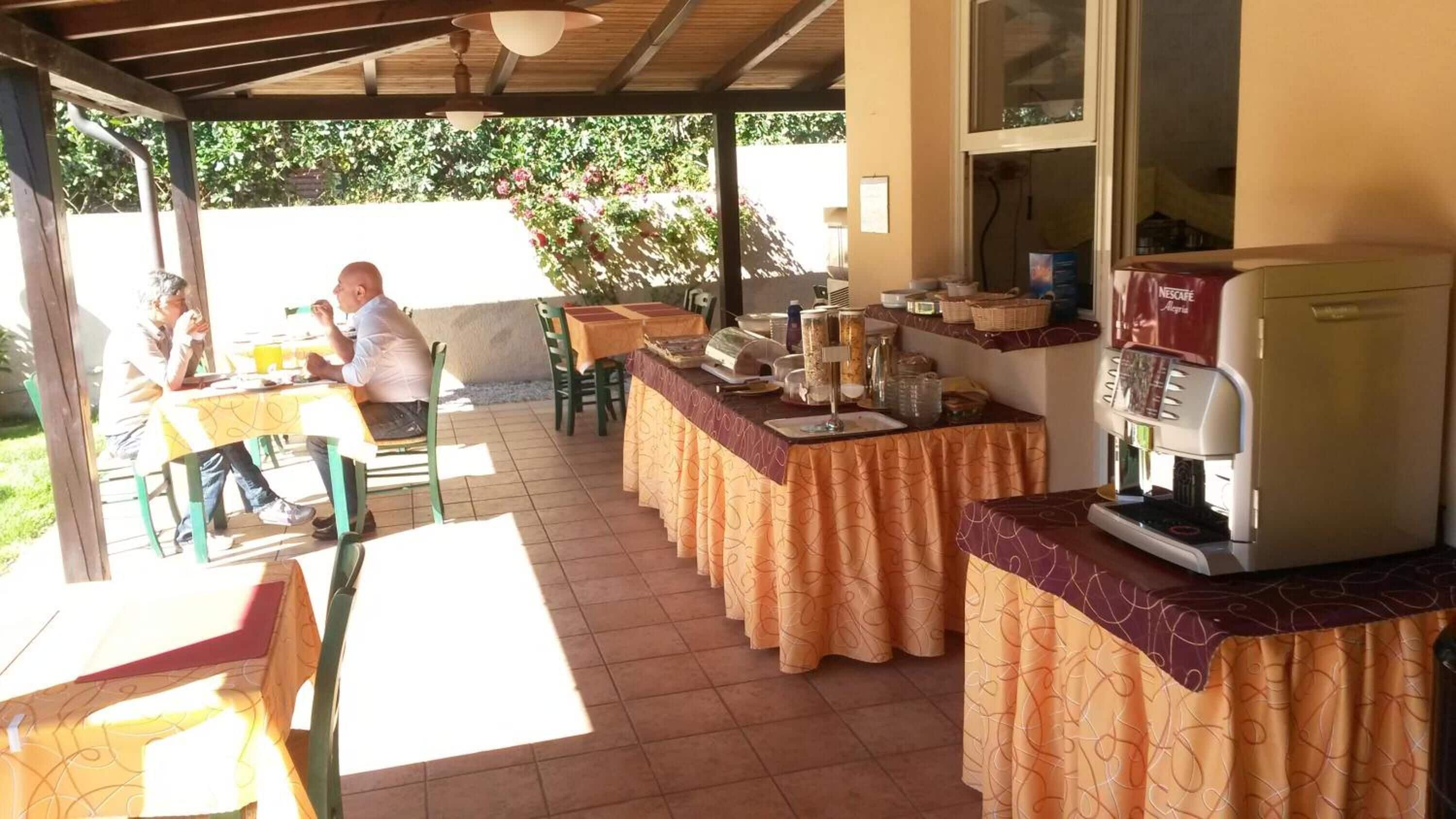 Un espacio comedor al aire libre techado, con mesas y sillas, una cafetera y un mostrador con varios artículos.