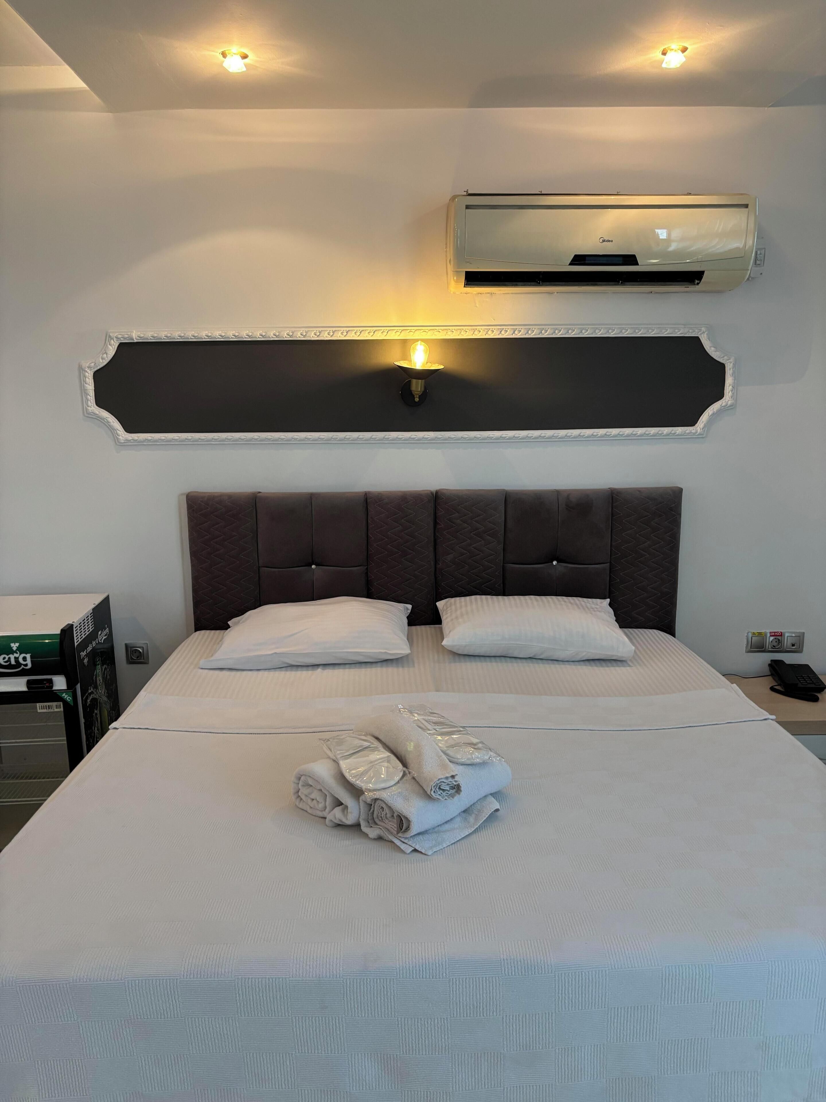 A hotel room with a large bed, two pillows, a folded towel, an air conditioner, and a wall-mounted light fixture.