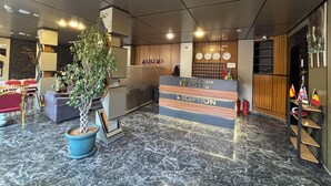 A hotel reception area with a reception desk, a potted plant, a clock, and flags on the wall.