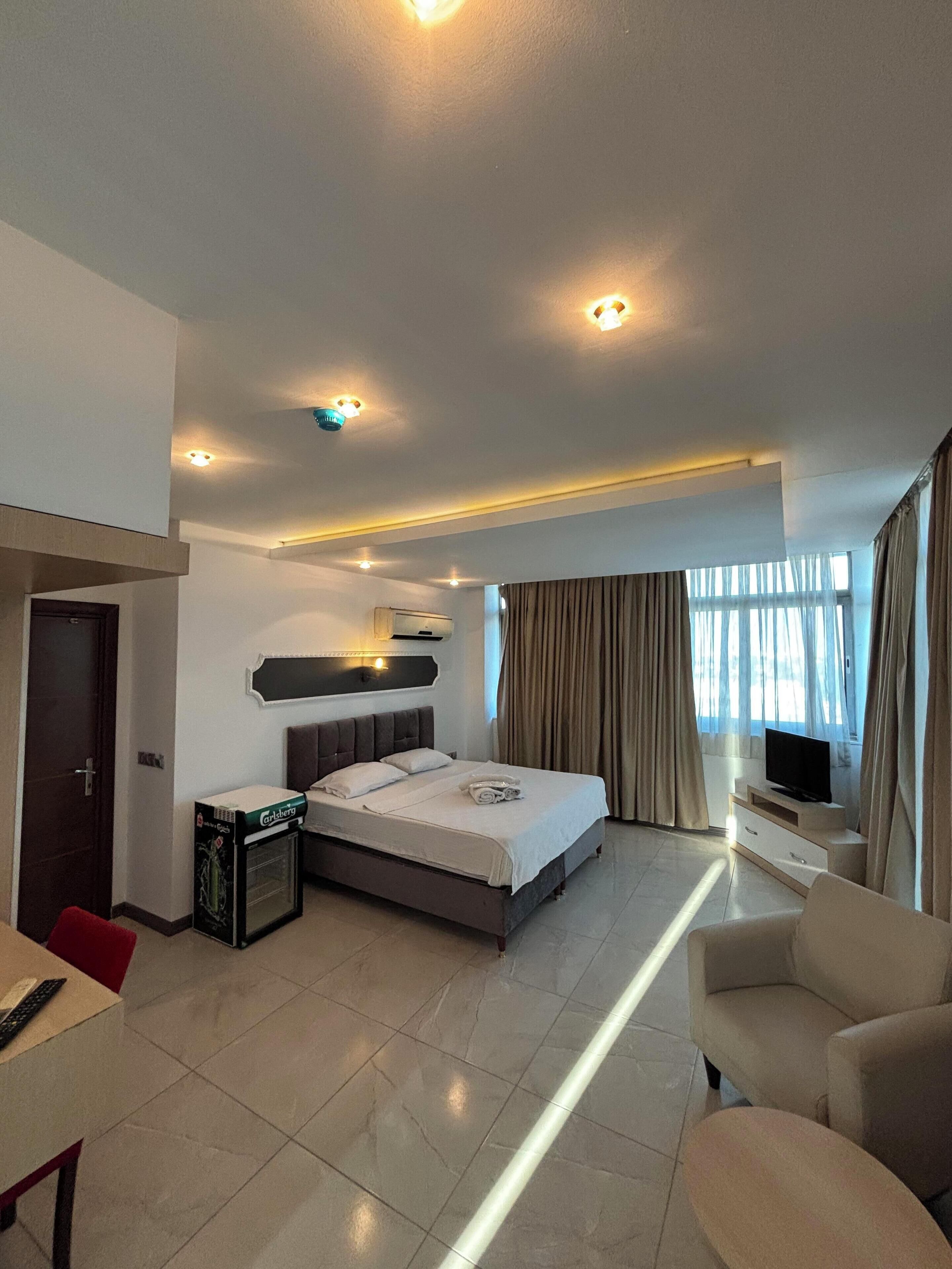 A modern hotel room with a large bed, a TV, a minibar, and a seating area with a chair and a small table.