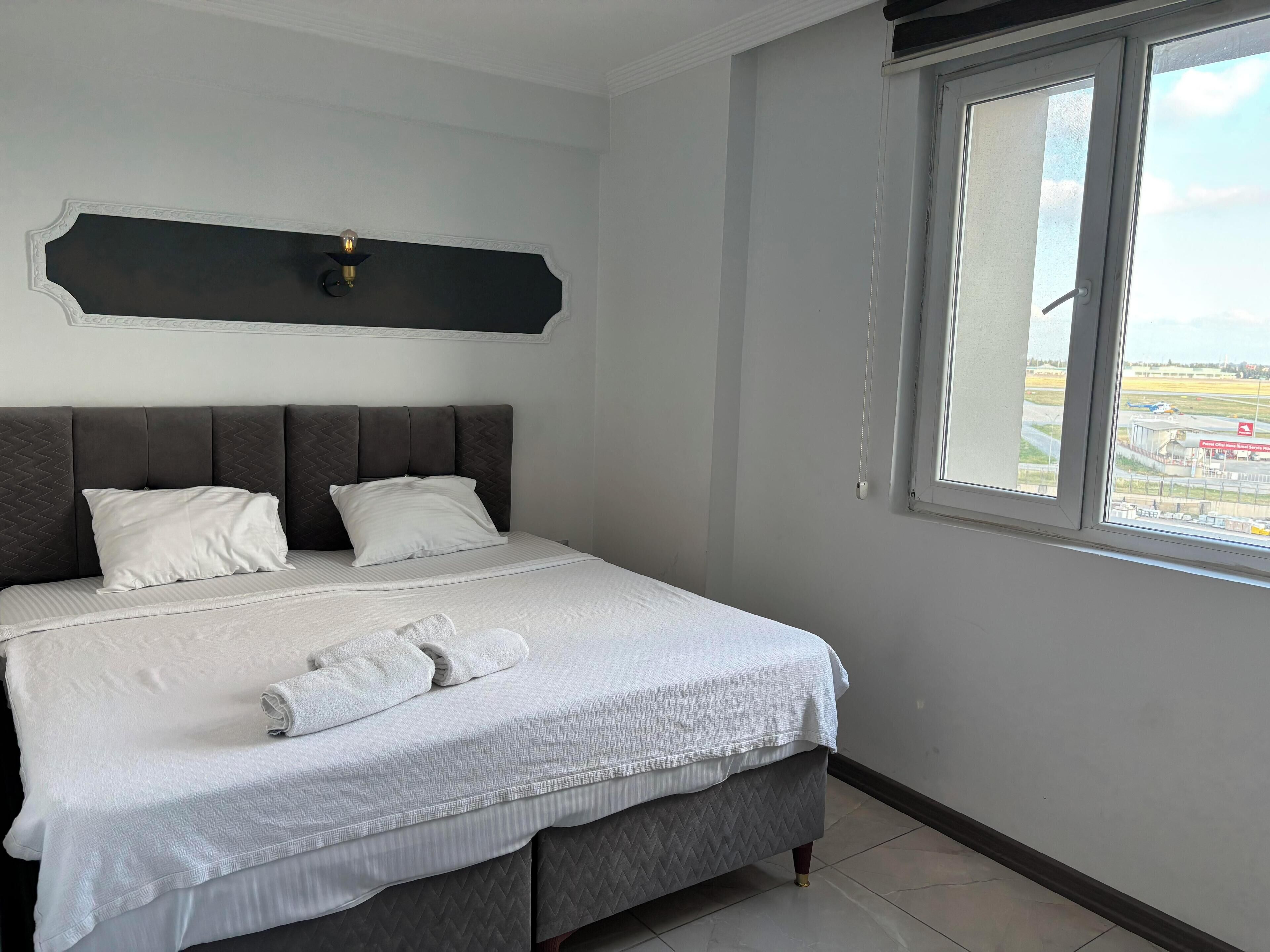 A neatly made bed with a grey headboard and white bedding, a window with a view of an airport, and a wall-mounted light fixture.