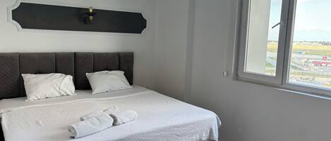 A neatly made bed with a grey headboard and white bedding, a window with a view of an airport, and a wall-mounted light fixture.