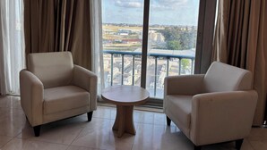 A room with two beige armchairs and a small round table, large windows with curtains, and a view of buildings and trees.