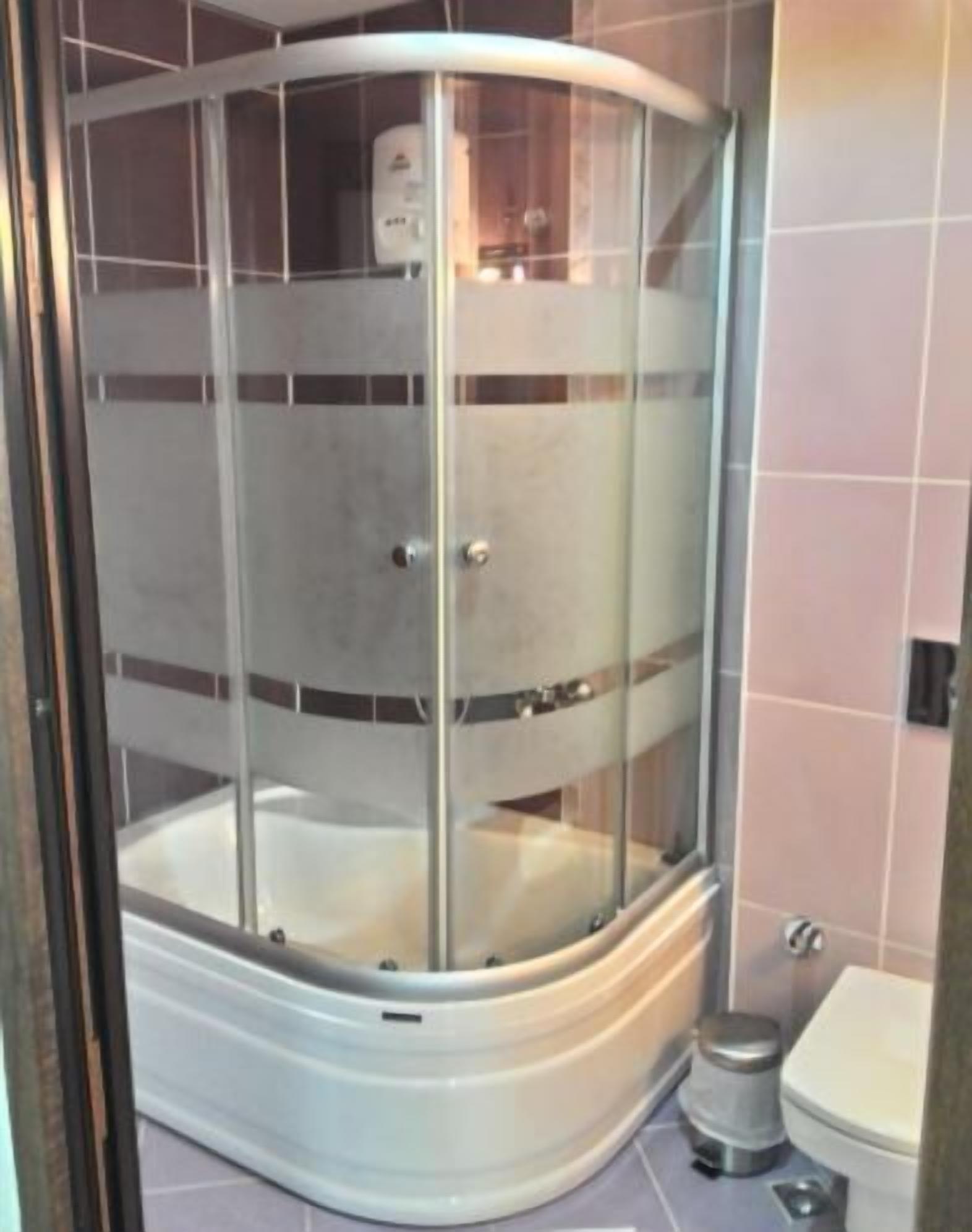 A glass-enclosed shower stall with a white base, adjacent to a toilet and a trash can.