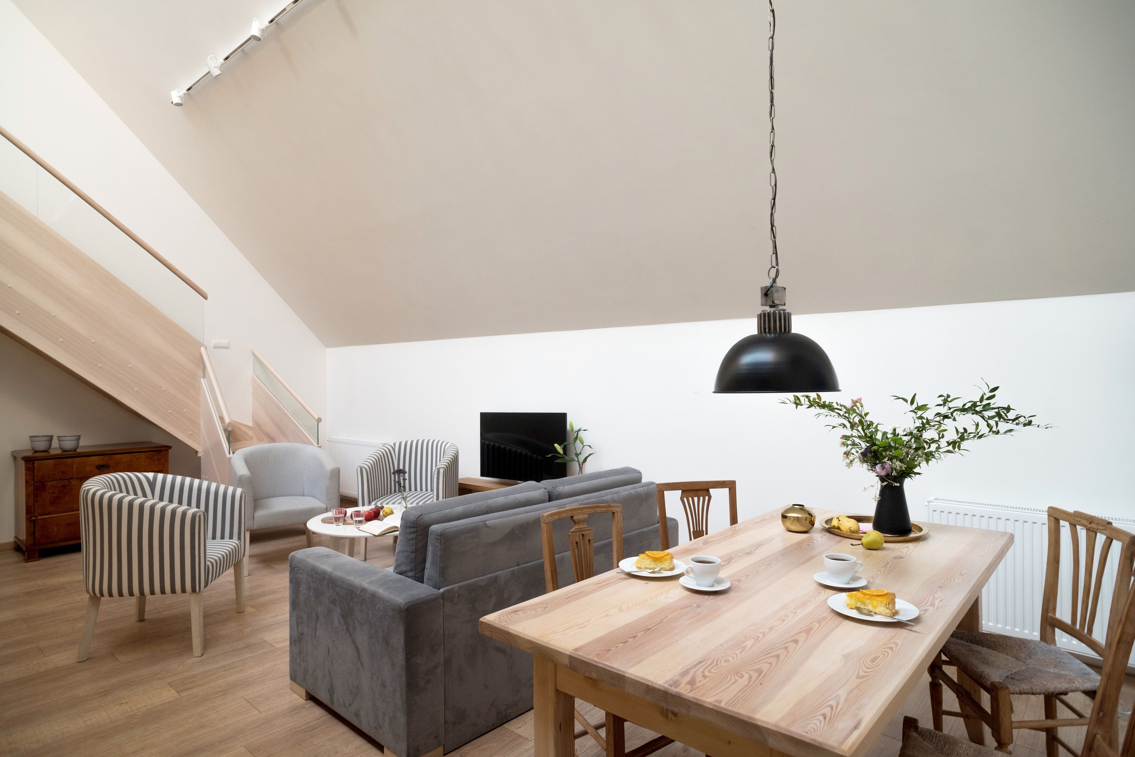 A modern living space with a wooden dining table, grey sofas, striped chairs, and a black pendant light.
