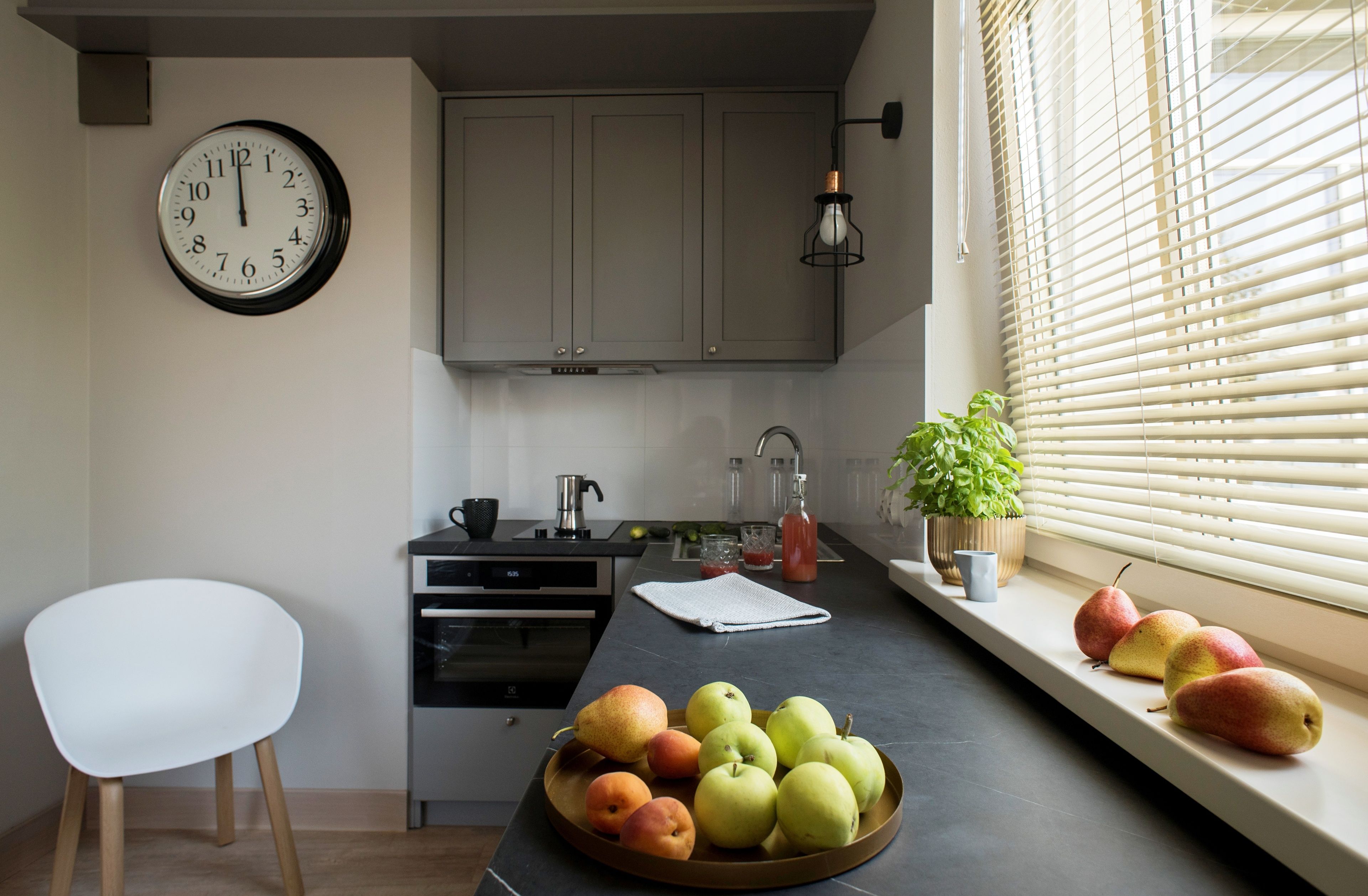 A kitchen with a clock, a sink, cabinets, a stove, a chair, and a fruit bowl.