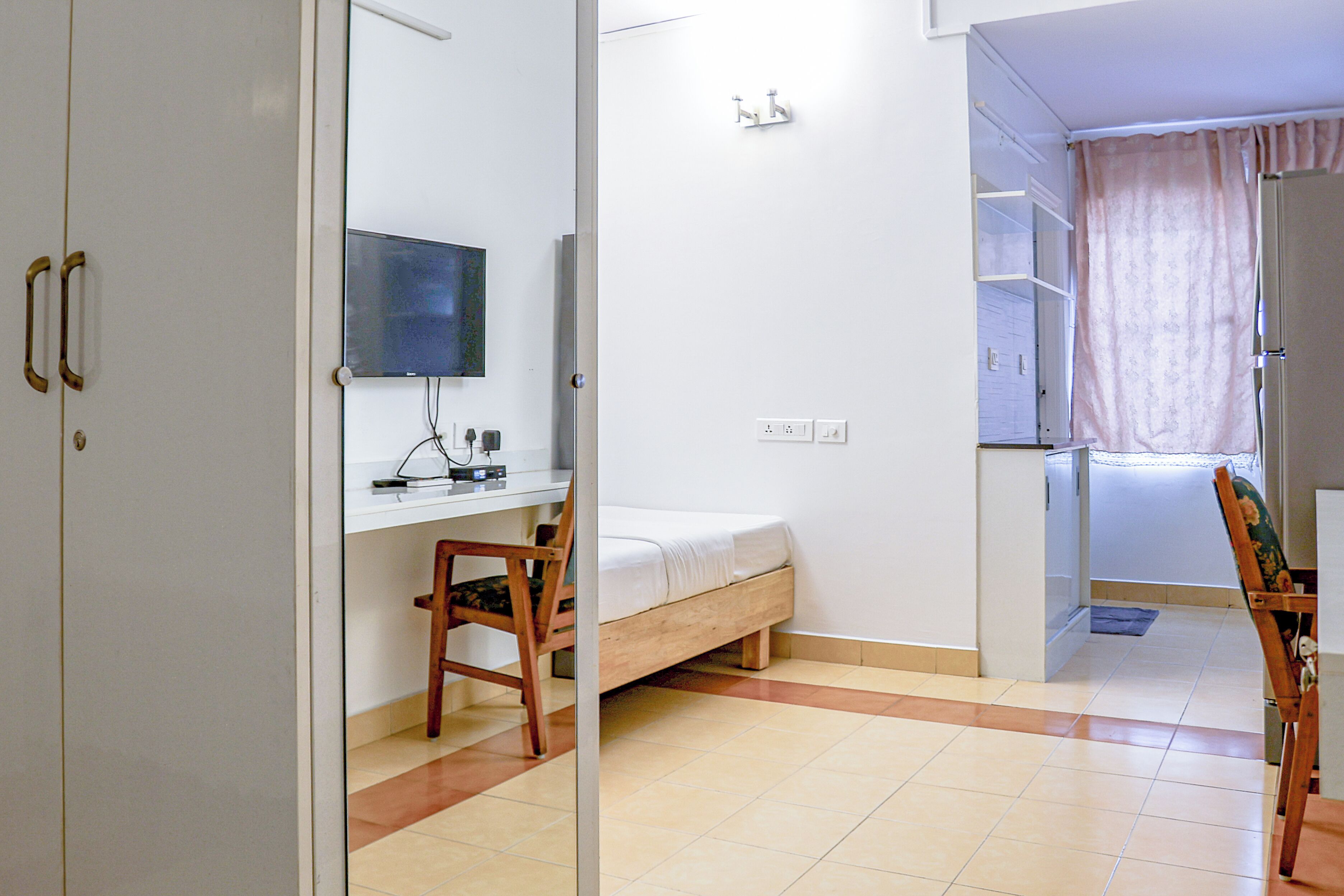 A bedroom with a bed, a desk, and a chair. There is a television on the wall and a window with curtains.