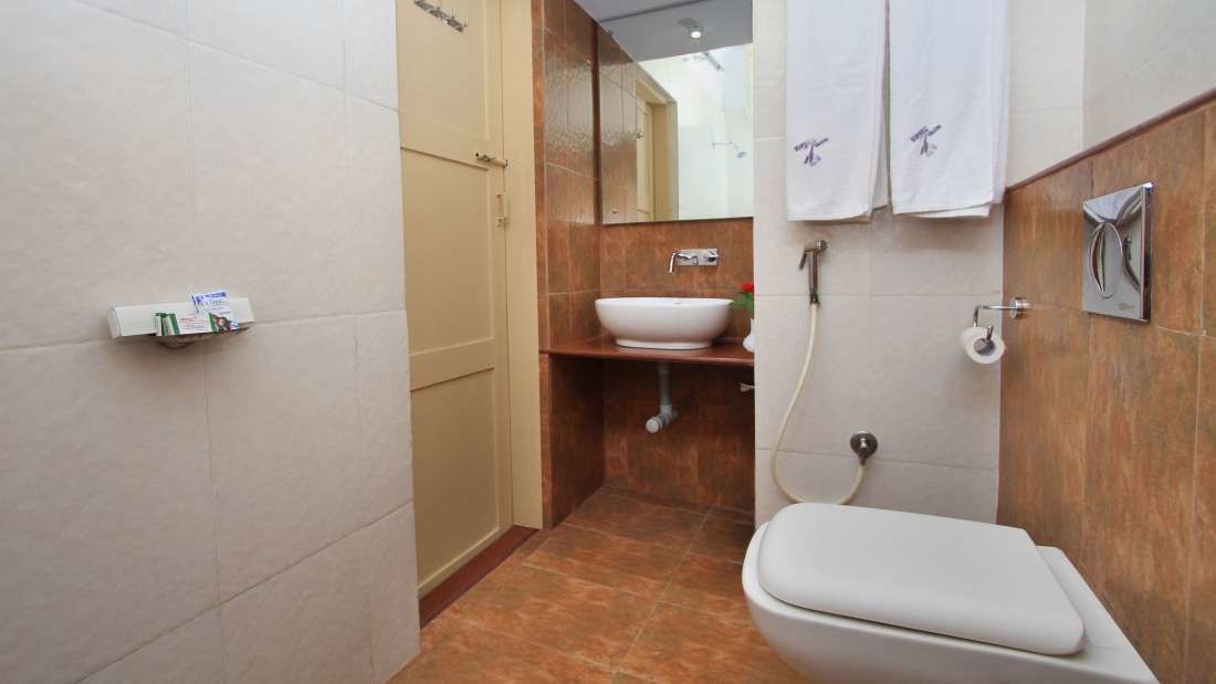A compact bathroom with a white toilet, a sink, and a shower. There is a towel rack with two towels and a small shelf with toiletries.