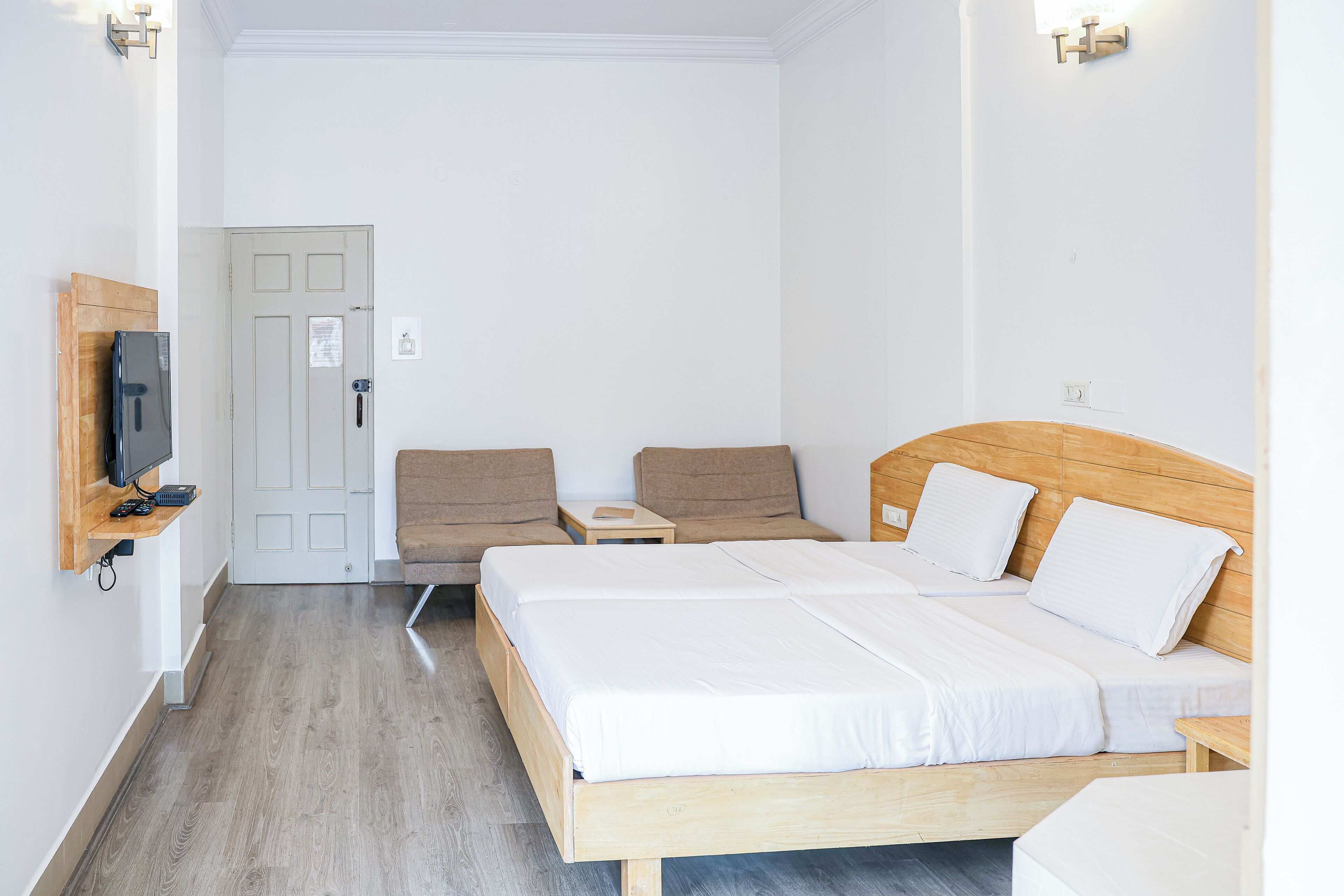 Una habitación de hotel con una cama de madera, dos sillas, una mesita y un televisor montado en la pared.
