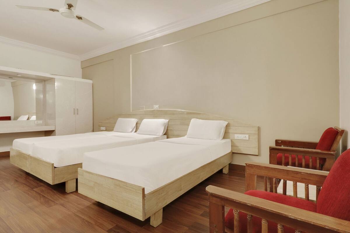 Une chambre d’hôtel avec deux lits simples, une chaise en bois, un miroir et un ventilateur de plafond.