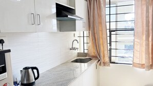 A kitchen with white cabinets, a stainless steel kettle, a sink, and a window with a curtain. - Hotel Vellara (Bengaluru)