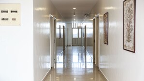 A long, well-lit hallway with doors on both sides, leading to a bright room with large windows. - Hotel Vellara (Bengaluru)