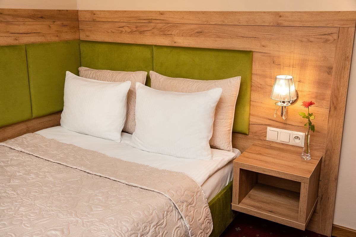 Uma cama bem arrumada com cabeceira verde, mesa de cabeceira com abajur e um vaso com uma flor.