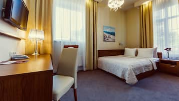 A hotel room with a bed, a desk, a chair, a TV, and a chandelier.