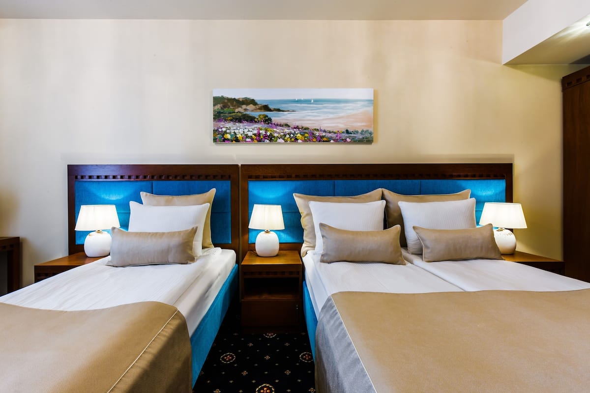 Quarto de hotel com duas camas, cada uma com cabeceira, abajures e uma pintura de uma praia acima das camas.
