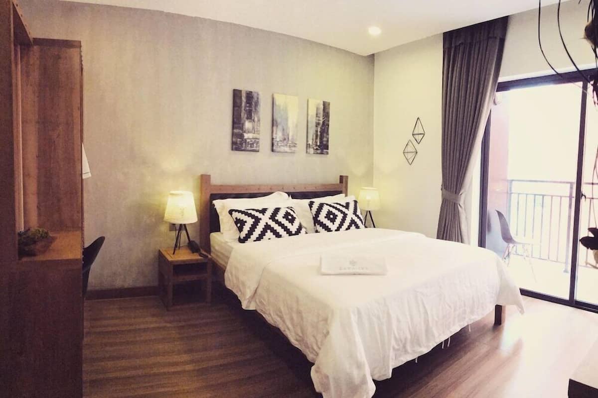 Habitación de hotel con cama, lámparas de noche, mesita de noche, una silla, ventana con cortinas y una obra de arte en la pared.