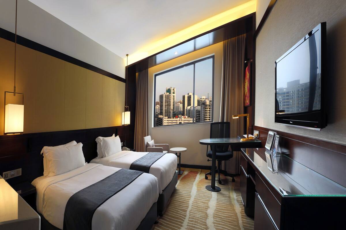 A hotel room with two beds, a desk, a chair, a TV, and a large window with a city view.