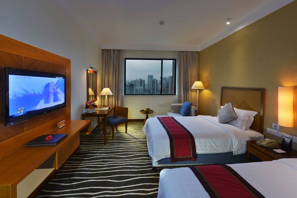 A hotel room with a television, two beds, a desk, a chair, a small table, and a window with a city view.