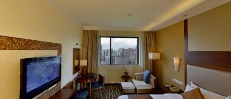 Une chambre d’hôtel comprenant un lit, une télévision, un bureau, une chaise, un canapé, une fenêtre donnant sur la ville et une table de chevet avec une lampe.