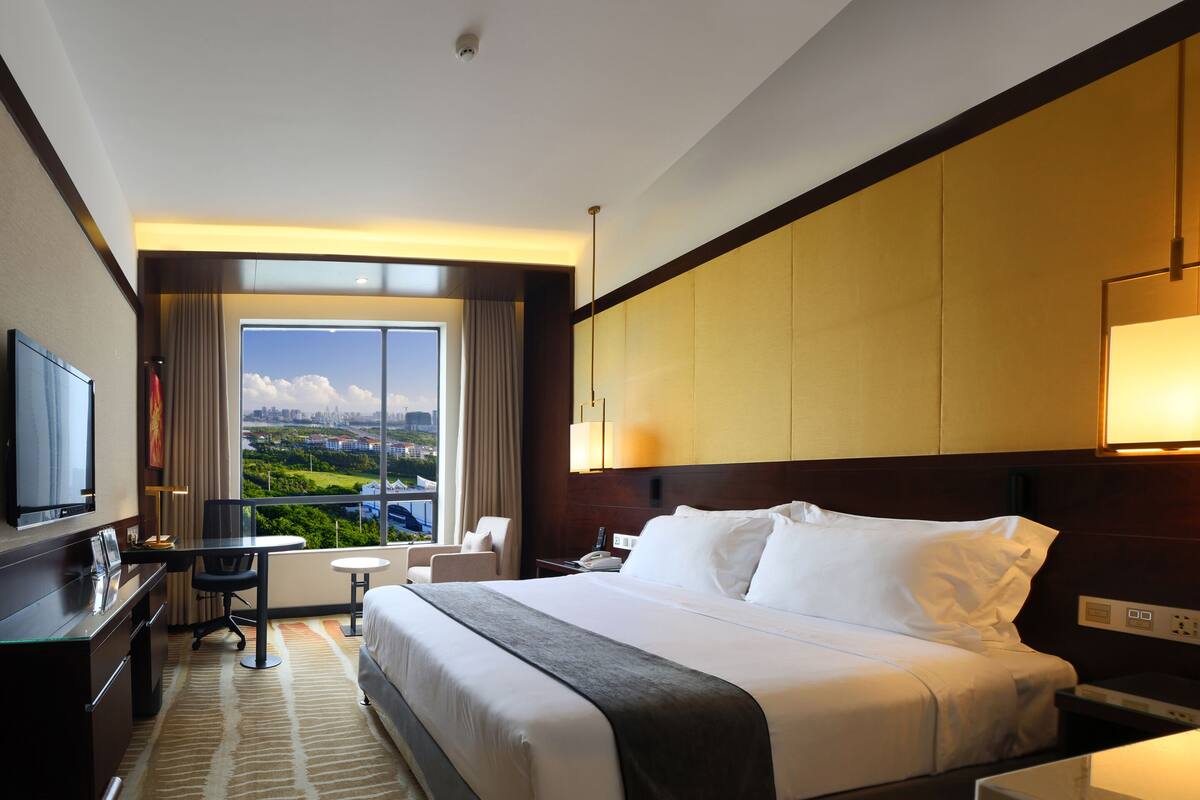 A hotel room with a large bed, a desk, a chair, a TV, and a view of the city through a large window.