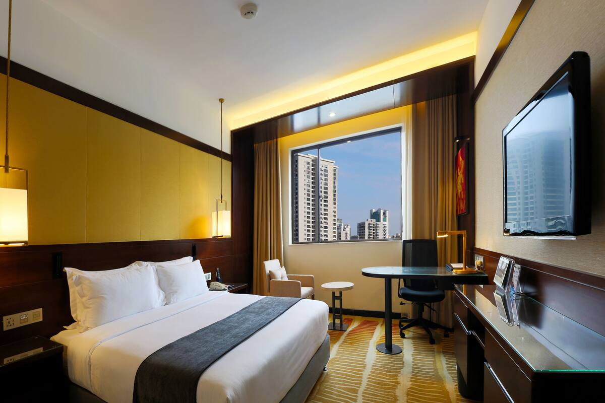 A hotel room with a large bed, a desk, a chair, a small table, and a TV mounted on the wall.