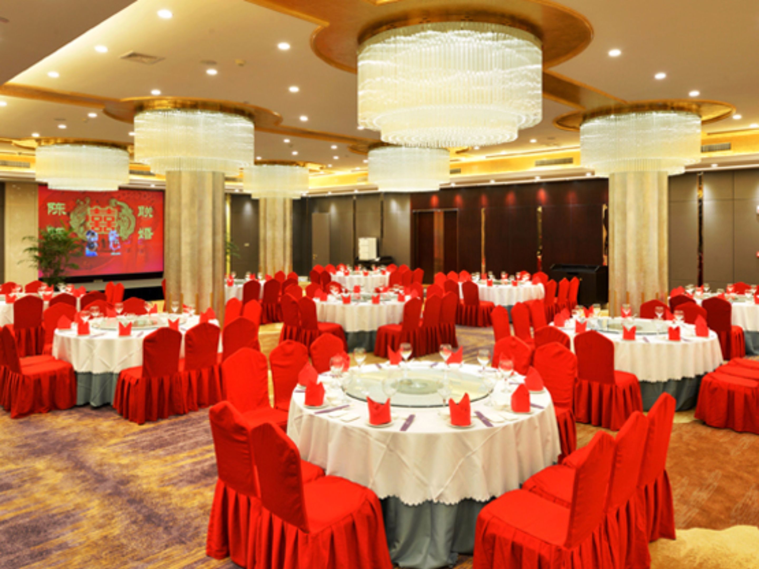 A banquet hall with round tables, red chairs, and chandeliers.