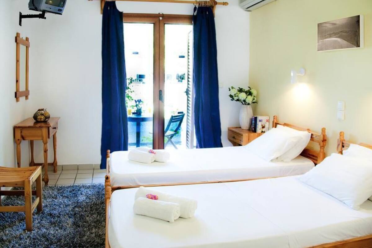 Habitación de hotel con dos camas individuales, una pequeña mesa de madera, una silla, un televisor montado en la pared y vista al exterior a través de un ventanal.