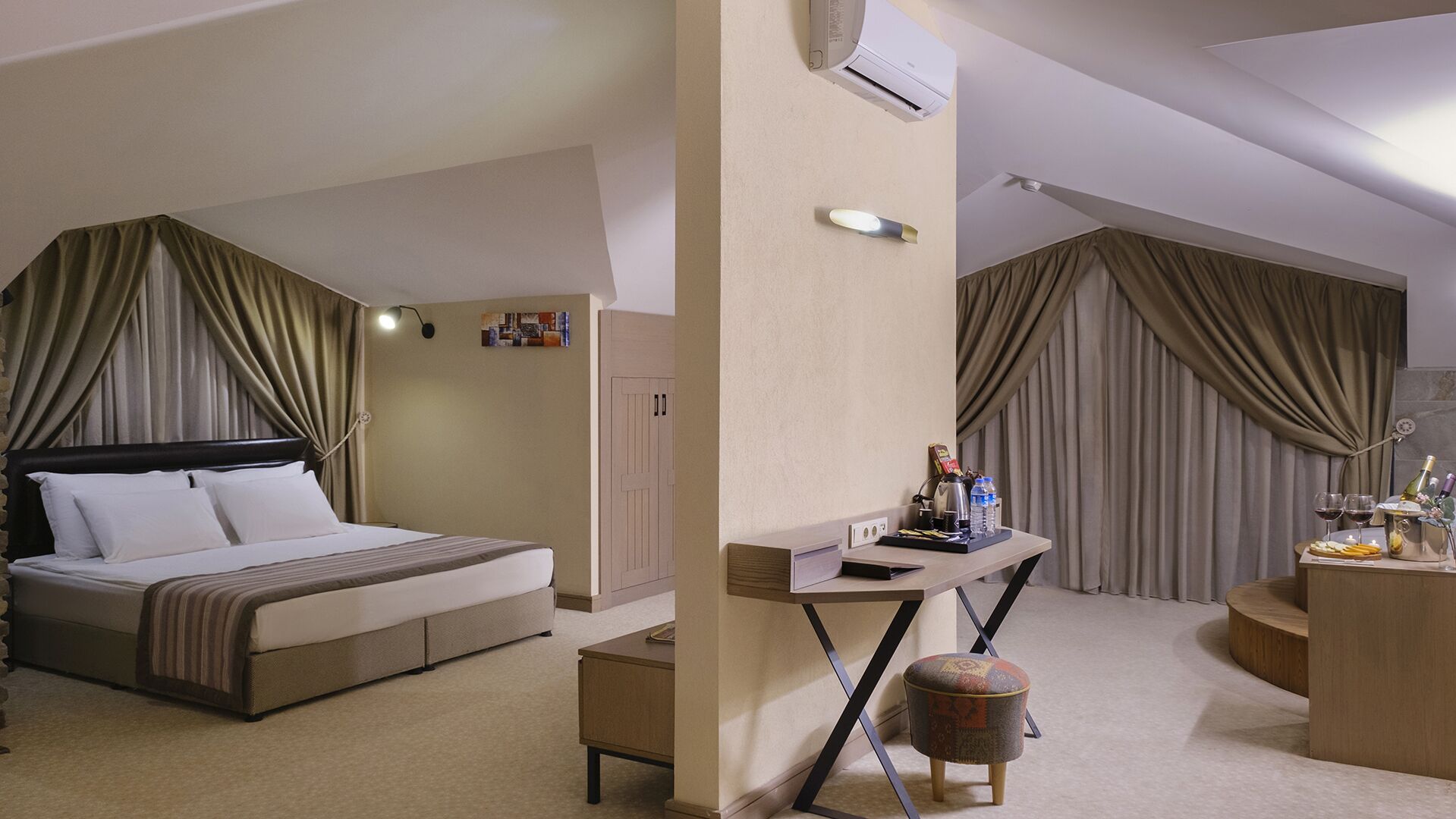 Habitación de hotel con cama, escritorio con silla, taburete pequeño y minibar con bebidas y aperitivos.