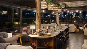 Un salón acogedor y bien iluminado con una larga barra de madera, asientos cómodos y grandes ventanales que ofrecen vista a la ciudad de noche.